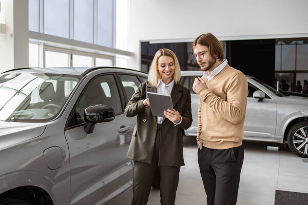 Уверенная блондинка-продавщица, объясняющая все особенности машины с помощью планшета красивому владельцу. Кавказский покупатель в автосалоне, женщина покупает транспорт, автодилер.
