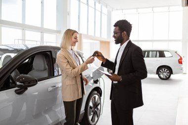 Takım elbiseli Afrikalı Amerikalı satış müdürü yeni elektrikli aracın yanında duran beyaz saçlı genç kadına araba anahtarı veriyor. Yeni araba konsepti seçmek ve satın almak.