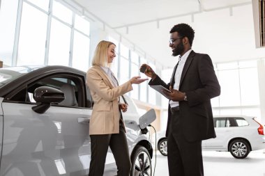 Takım elbiseli Afrikalı Amerikalı satış müdürü yeni elektrikli aracın yanında duran beyaz saçlı genç kadına araba anahtarı veriyor. Yeni araba konsepti seçmek ve satın almak.