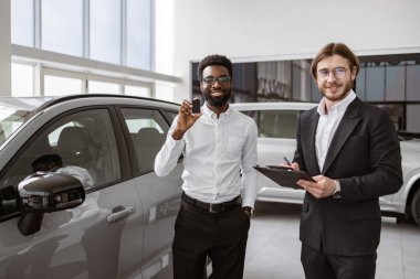 Mutlu iş adamının elinde anahtarlar var. Parlak bayilikte takım elbiseli mutlu bir satıcının yanında yeni bir araba almış. Takım elbiseli beyaz adam, modern SUV 'yi Afrikalı iş adamına sattı..