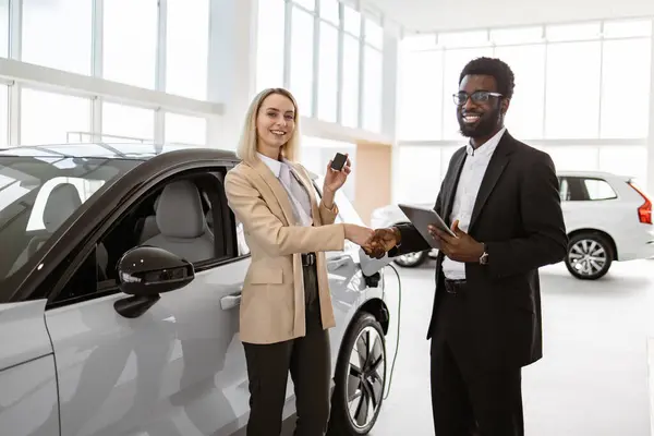 ハンサムなアフリカ人男性は 車のバイヤー 若い白人ビジネスレディと握手し 明るいディーラーに立っています 車のショールーム 新しい車を購入する女性 ストック画像