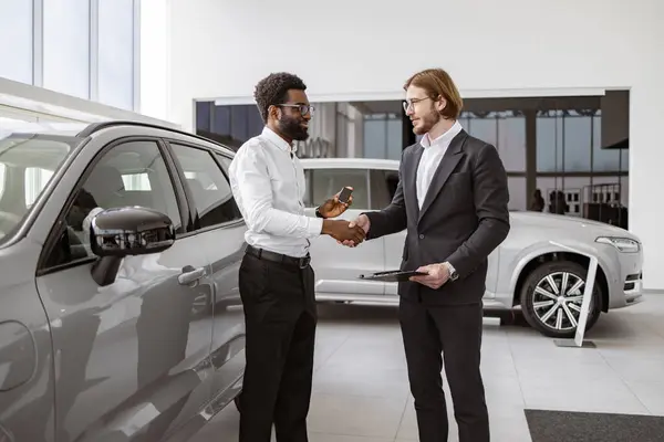 ハンサムなアフリカ人男性は 車の売り手 若い白人セールスマンと握手し 明るいディーラーの近くに立っています 車のショールーム 新しい車を購入する男 ロイヤリティフリーのストック写真