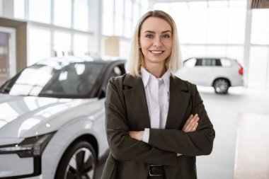 Parlak ve geniş sergi salonunda kolları çapraz duran beyaz takım elbiseli güzel araba satıcısı portresi. Araba galerisindeki profesyonel kadın satış elemanı..