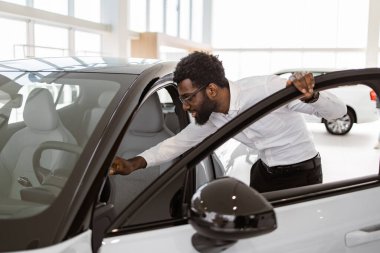 Genç Afrikalı erkek araba seçiyor, galeride yeni bir araba almak istiyor. Mutlu sakallı adam müşteri, beyaz gömlek giyiyor, galeride arabanın içini inceliyor..