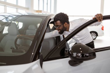 Genç Afrikalı erkek araba seçiyor, galeride yeni bir araba almak istiyor. Mutlu sakallı adam müşteri, beyaz gömlek giyiyor, galeride arabanın içini inceliyor..