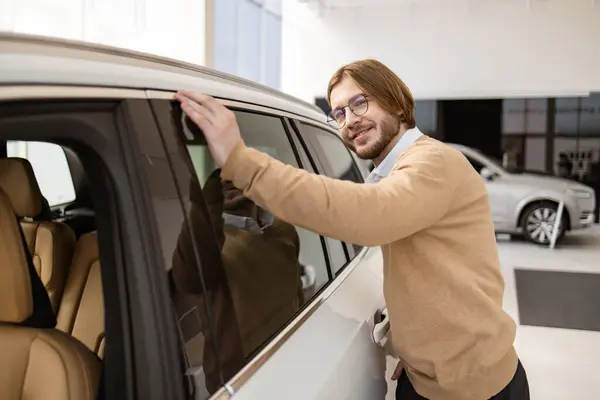 ハッピーで楽しい男性の顧客バイヤー クライアント 黄色いセーターを着て 下の窓から車の内部を見ています 自動車を選ぶひげ付きの若い男性は 車のショールームで新しい自動車を購入したいです ロイヤリティフリーのストック写真