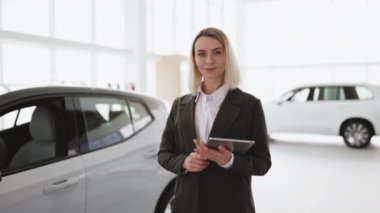Yeni araba galerisindeki kameraya bakarken elinde tablet olan gülümseyen satıcı kadın. Profesyonel araba satıcısı gururla oto galeride poz veriyor, kameraya gülümsüyor, elinde tablet var..