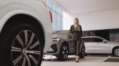 Dijital tabletli, gülümseyen ve kameraya bakan profesyonel beyaz kadın satıcısı. Parlak araba galerisinde modern otomobilin yanında duran genç satış elemanı..