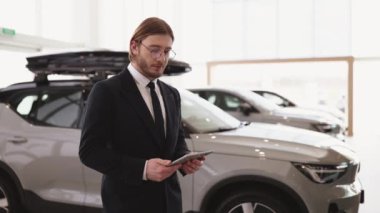 Gülümseyen beyaz satıcı kameraya bakıyor ve sergi salonundaki yeni araba modellerini işaret ediyor. Araba galerisinde gururla poz veren takım elbiseli ve gözlüklü profesyonel sakallı bir araba satıcısı..