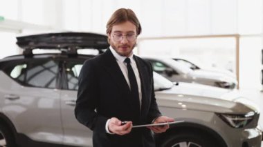 Kendine güvenen satıcı, galerideki yeni arabanın anahtarını müşteriye veriyor. Araba satıcısı neşeyle gülümsüyor, anahtarı tutuyor, satılık yeni araçların önünde duruyor. Teklif, indirim kavramı.