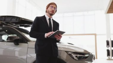 Gözlüklü ve takım elbiseli genç araba satıcısı dijital tablet tutuyor. Kendine güvenen sakallı bir adam modern aletlerle yeni bir araba satın almak için emir verir..