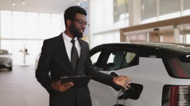 İlerici müşteriler için çevre dostu araba alma kavramı. Genç sakallı Afrikalı satıcı, panoramik pencereli modern sergi salonunda elektrikli araba satıyor..