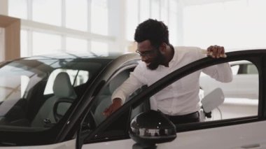 Genç Afrikalı erkek araba seçiyor, galeride yeni bir araba almak istiyor. Mutlu sakallı adam müşteri, beyaz gömlek giyiyor, galeride arabanın içini inceliyor..