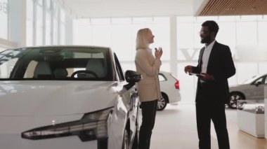 Takım elbiseli Afrikalı Amerikalı satış müdürü yeni elektrikli aracın yanında duran beyaz saçlı genç kadına araba anahtarı veriyor. Yeni araba konsepti seçmek ve satın almak.