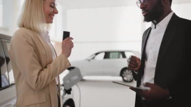 Takım elbiseli Afrikalı Amerikalı satış müdürü yeni elektrikli aracın yanında duran beyaz saçlı genç kadına araba anahtarı veriyor. Yeni araba konsepti seçmek ve satın almak.