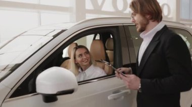 Yeni modern SUV arabasının içinde oturan mutlu beyaz kadın müşteri. Kendine güvenen erkek tezgahtar, aydınlık galeriden araba aldıktan sonra arabanın anahtarlarını veriyor..