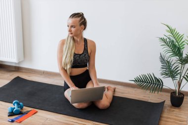 Spor kıyafetli genç bir kadın aydınlık odada yoga minderinde oturuyor. Yanında spor malzemeleri olan bir dizüstü bilgisayar kullanıyor. Mutlu kadın meşgul ve memnun görünüyor, teknolojinin ve sağlıklı yaşam tarzlarının karışımını vurguluyor..