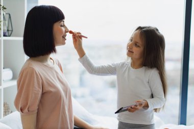 Kafkas kızının genç annelerinin yüzüne pudra sürerken yakın plan portresi. Çekici annesine makyaj yapan sevimli küçük kız, büyük pencerenin yanında yatağında oturuyor..