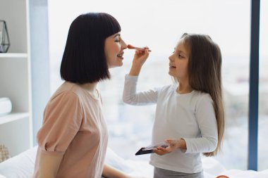Kafkas kızının genç annelerinin yüzüne pudra sürerken yakın plan portresi. Çekici annesine makyaj yapan sevimli küçük kız, büyük pencerenin yanında yatağında oturuyor..