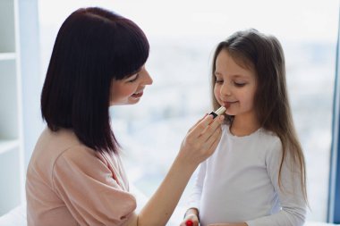 Yatak odasında oturan küçük kız annesine makyaj yapıyor. Mutlu anne ve kız hafta sonu birlikte eğleniyorlar. Kız, güzel annesine dudak boyuyor. Mutlu sevgi dolu aile.