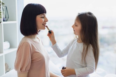 Mutlu anne ve kız hafta sonu birlikte eğleniyorlar. Yatak odasında oturan küçük kız annesine makyaj yapıyor. Kız, güzel annesine dudak boyuyor. Mutlu sevgi dolu aile.