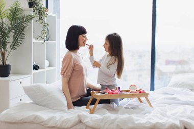 Mutlu anne ve kız hafta sonu birlikte eğleniyorlar. Yatak odasında oturan küçük kız annesine makyaj yapıyor. Kız, güzel annesine dudak boyuyor. Mutlu sevgi dolu aile.