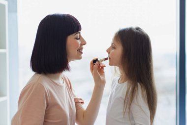 Mutlu anne ve kız hafta sonu birlikte eğleniyorlar. Yatak odasında oturan küçük kız annesine makyaj yapıyor. Kız, güzel annesine dudak boyuyor. Mutlu sevgi dolu aile.