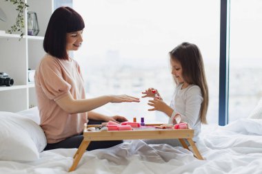 Şirin küçük kız anne ellerine oje sürerek oje sürmeyi öğreniyor. Mutlu anne ve kız yataklarında oturup manikür yapıyor, vücutlarıyla ilgileniyorlar..