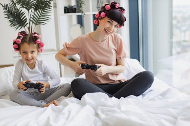 Hafta sonları evde eğlenceli aile zamanları. Kafkas anne ve küçük kızı video oyunu oynarken saçlarını kıvırıyor, panoramik pencerenin yanında yatıyorlar..