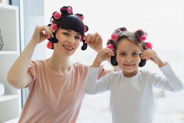Gülümseyen anne ve kızın bigudilerle portresi. Yatak odasındaki pencerenin arka planında ellerinde göz bantları var..