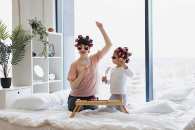 Evdeki güzellik tedavileri sırasında tarakları mikrofon olarak kullanarak şarkı söyleyen neşeli anne ve kız. Kafkasyalı genç bir kadın ve küçük bir kız saçları kıvırıyor ve göz bantları takıyor..