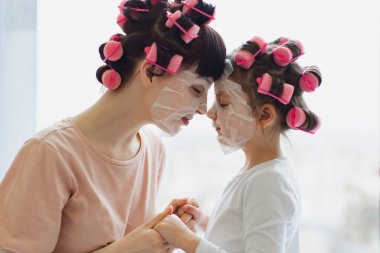 Anne ve kızı saçlarını kıvırıp nemlendirici yüz maskesi takarken alınlarına dokunuyorlar. Deri ve saç bakımı ve aile ilişkileri kavramı.