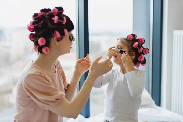 Happy Caucasian family spends free time having cosmetic procedures at home on bedroom. Young mother and cute little girl doing hair perm and using eye patches at weekend.