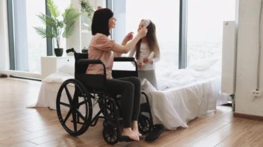 Genç anne tekerlekli sandalye kullanıcısı küçük kızının saçını tarıyor yatak odasındaki panoramik pencerenin yanında oturuyor. Mutlu Kafkas ailesi hafta sonu sabahları eğleniyor..