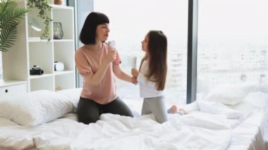 Genç anne, yatakta otururken mikrofon yerine tarak kullanarak sevimli kızıyla şarkı söylüyor. Mutlu Kafkas ailesi hafta sonu sabahı eğleniyor..
