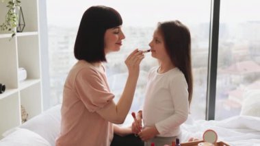 Yatak odasında oturan küçük kız annesine makyaj yapıyor. Mutlu anne ve kız hafta sonu birlikte eğleniyorlar. Kız, güzel annesine dudak boyuyor. Mutlu sevgi dolu aile.