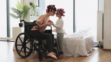 Mutlu beyaz kadın tekerlekli sandalye kullanıcısı ve küçük kız evde, yatak odasında kozmetik ameliyatlar yapıyor. Genç bir kadın ve güzel bir kız saç perması yapıyor ve bantları yapıştırıyor..