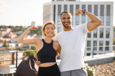 Sağlıklı yaşam tarzını geliştirirken kaslarını gösteren gülümseyen atletik erkek ve kadın. Spor giyimli genç bir çift çatıdaki kentsel binaların arka planında poz veriyor.