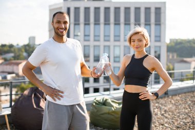 Atletik adam ve zayıf kadın sabah koşularından sonra dinleniyorlar. Modern evin çatısında başarılı bir çalışmadan sonra su şişelerini tokuşturan mutlu çift..