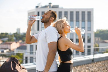 Atletik formda dişiler sırtı kaslı sporcuya dönük durur ve su içer. Şehir geçmişi olan yüksek binaların terasında beden eğitimi alan genç bir çift..