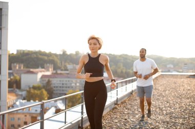Sabahları siyah tişörtlü genç bir kadın beyaz tişörtlü bir erkekle şehir geçmişine karşı koşuyor. Sağlıklı yaşam tarzı kavramı ve günün olumlu başlangıcı.