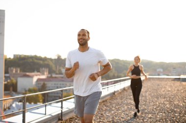 Beyaz tişörtlü ve şortlu gülümseyen atletik erkek sabahları modern yüksek binaların terasında koşuyor. Genç bayan, bulanık arka planda koşuyor..