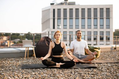 Spor giyimli genç bir çift modern evin çatısında lotus pozisyonunda minderlerin üzerinde oturuyor. Atletik bir erkek ve esnek bir kadın sabah şehir geçmişi üzerine meditasyon yapıyorlar..