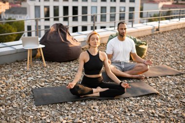 Spor giyimli genç bir çift modern evin çatısında lotus pozisyonunda minderlerin üzerinde oturuyor. Atletik bir erkek ve esnek bir kadın sabah şehir geçmişi üzerine meditasyon yapıyorlar..