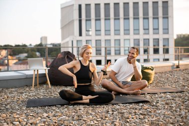 Atletik adam ve zayıf kadın sabah ısınması sırasında bacak bacak üstüne atarak oturuyorlar. Genç mutlu çift yoga dersinden sonra modern evin çatısında dans ediyor..