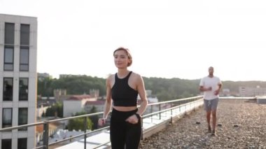 Siyah tişörtlü zayıf mutlu kadın, şehir arka planındaki modern binanın çatısında sabah ısınması sırasında beyaz tişörtlü sporcu adamla koşu yarışını kazanıyor. Sağlıklı yaşam tarzı kavramı.