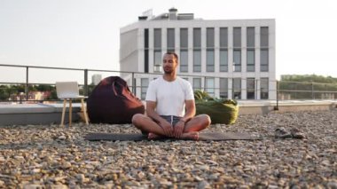 Genç atletik adam gün batımında şehir geçmişinde yoga yapıyor. Yakışıklı erkek Lotus pozisyonunda meditasyon yaparken modern evin çatısında oturuyor..