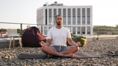 Yakışıklı erkek, modern evin çatısında otururken lotus pozisyonunda meditasyon yapıyor. Genç atletik adam gün batımında şehir geçmişinde yoga yapıyor..
