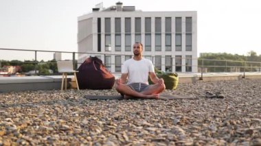 Yakışıklı erkek, modern evin çatısında otururken lotus pozisyonunda meditasyon yapıyor. Genç atletik adam gün batımında şehir geçmişinde yoga yapıyor..