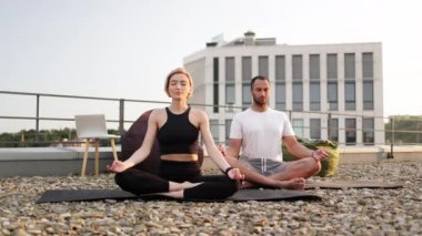 Atletik bir adam ve esnek bir kadın sabah şehir geçmişi üzerine meditasyon yapıyorlar. Spor giyimli genç bir çift modern evin çatısında lotus pozisyonunda minderlerin üzerinde oturuyorlar..